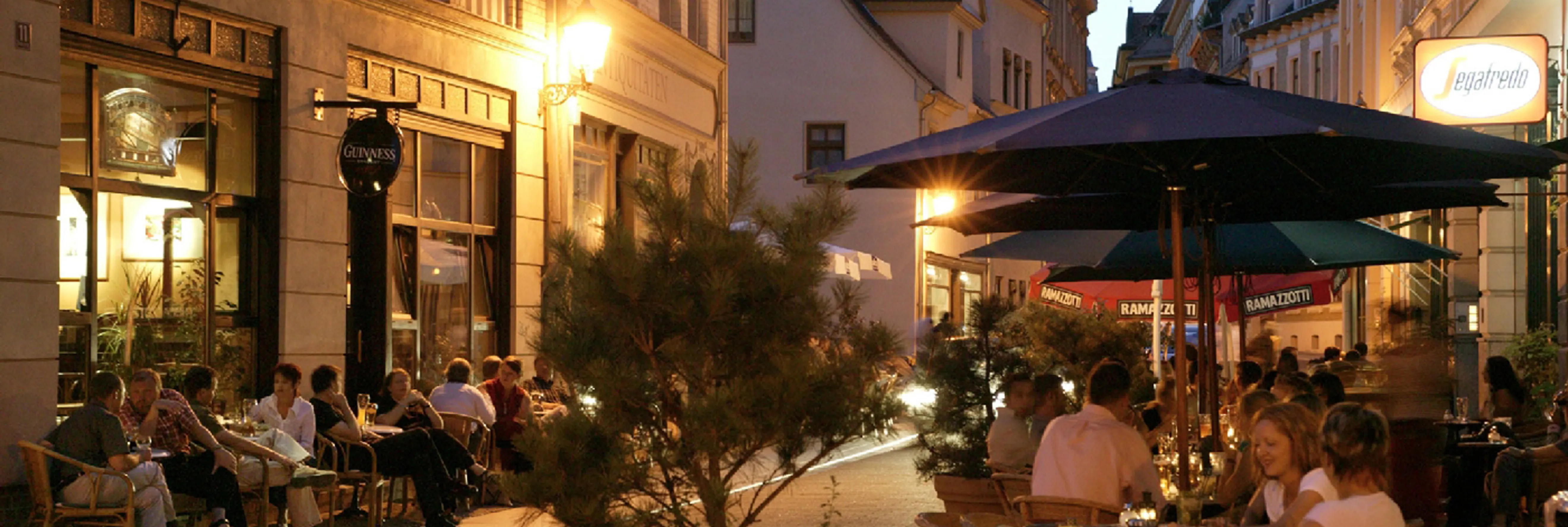Eine belebte Straße am Abend ist zu sehen: In den Restaurants und Bars sitzen viele Menschen im Außenbereich.